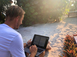 Ein Mann sitzt am Strand und arbeitet an einem Laptop, während die Sonne durch die Blätter schein
